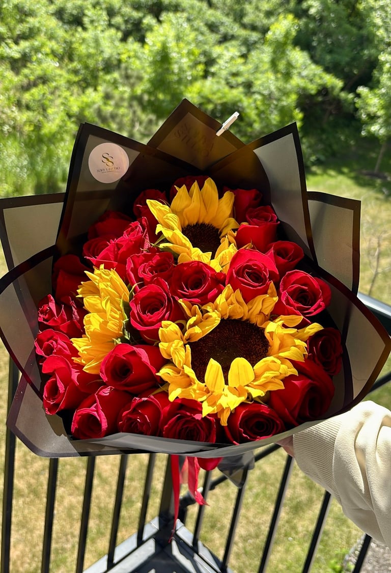 Bouquet of red roses and yellow sunflowers wrapped in black paper, held outdoors with green foliage background