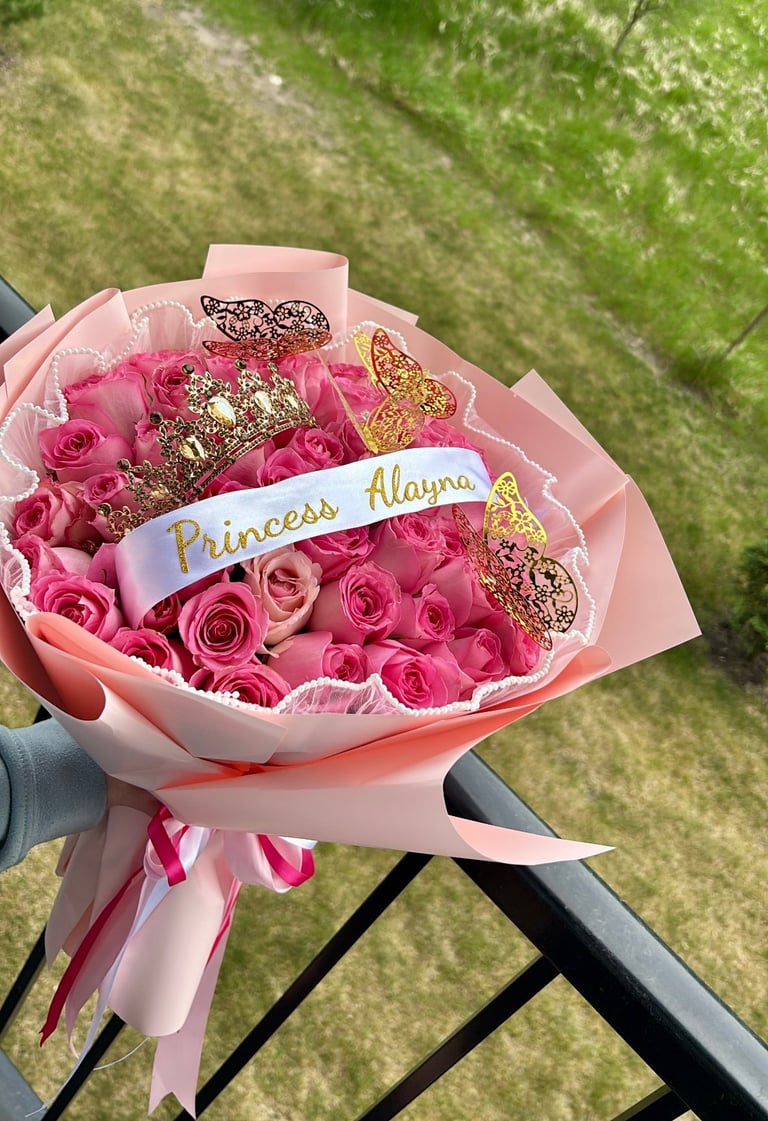 Pink rose bouquet with gold butterfly decorations and Princess Alayna ribbon, held outdoors on grass