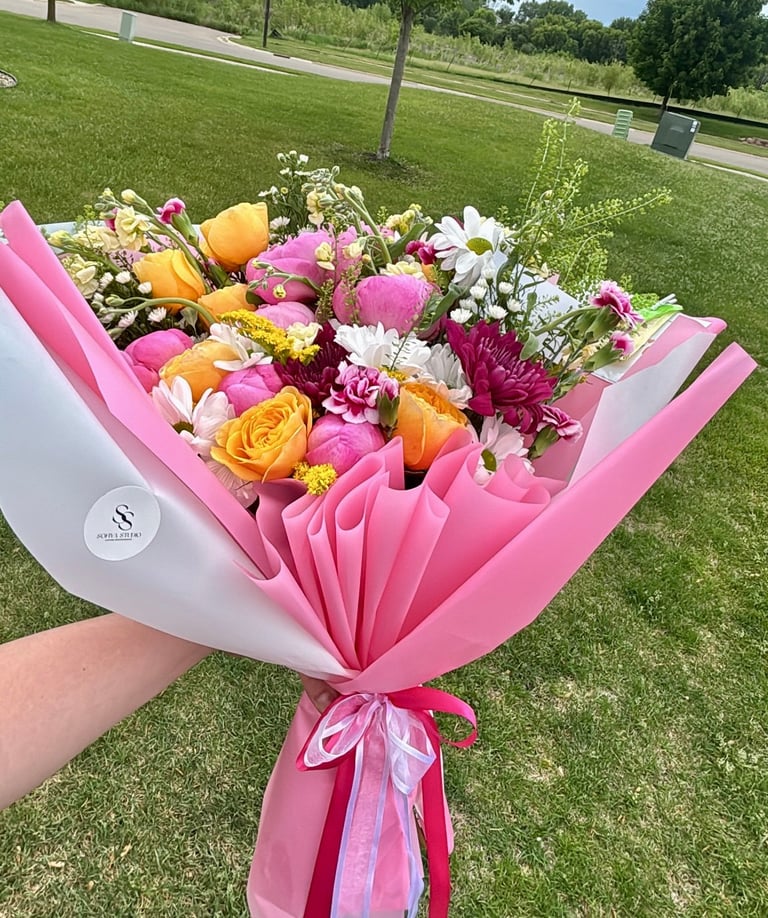 Hand holding a colorful bouquet of pink, orange, and white flowers wrapped in pink and white paper with a ribbon bow, displayed outdoors on a green lawn