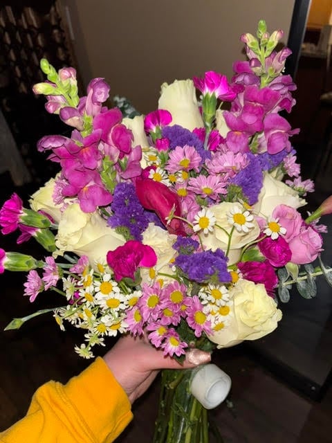 Person in yellow sleeve holding vibrant bouquet of magenta snapdragons, white roses, purple statice, and white daisies