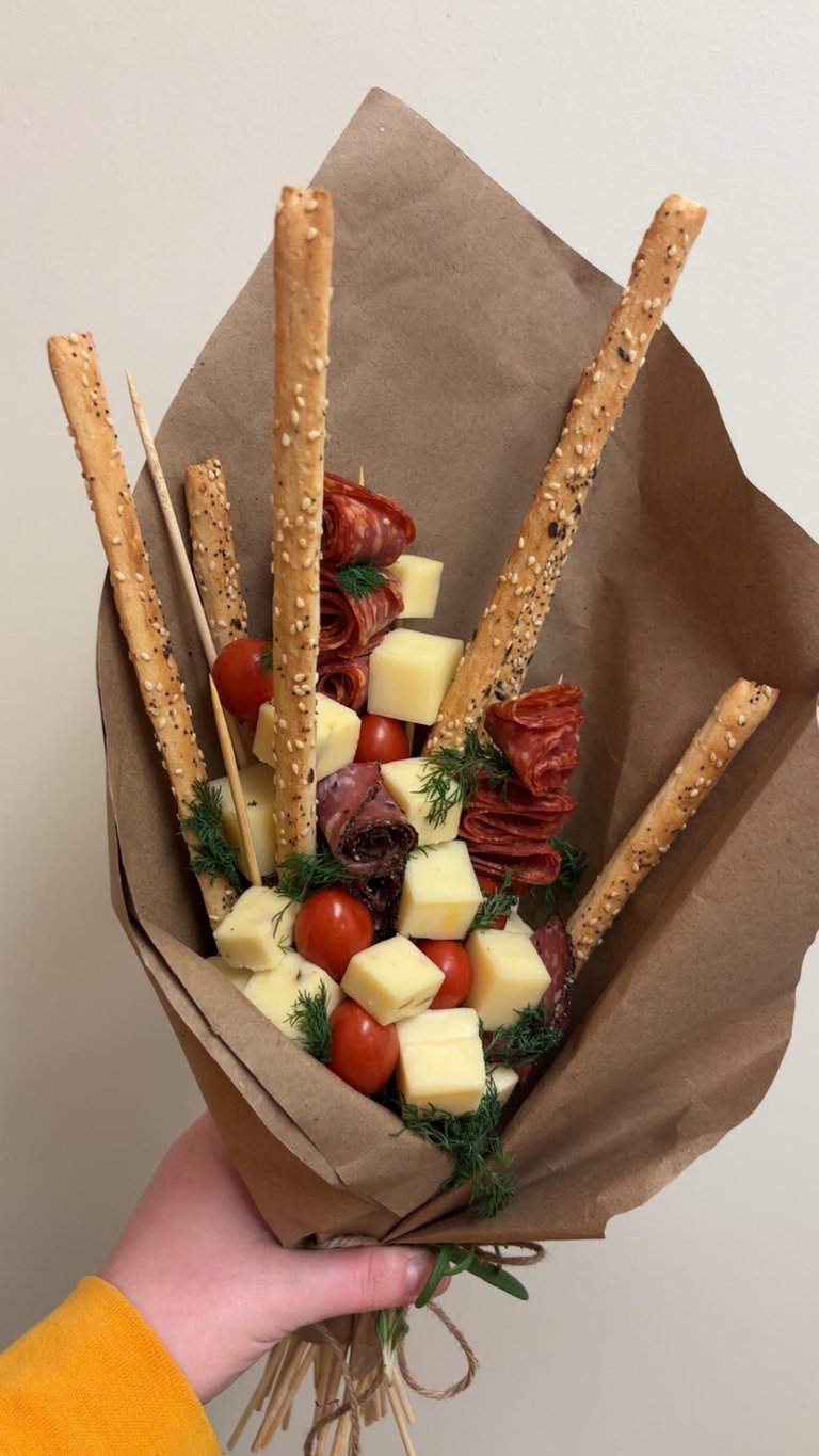 Hand holding a charcuterie bouquet wrapped in brown paper with breadsticks, cured meats, cheese cubes, cherry tomatoes, and fresh dill