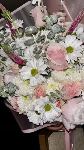 A fresh flower bouquet with white daisies, pink roses, cream chrysanthemums, and dusty miller foliage tied with pink ribbon