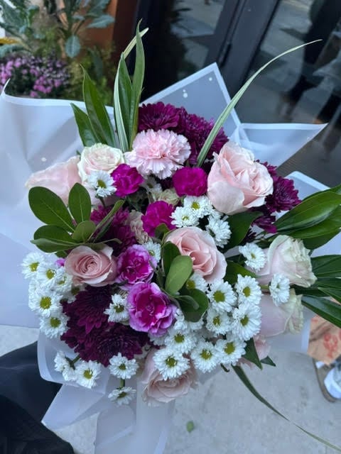 Elegant floral bouquet with magenta dahlias, pink roses, white daisies, greenery, and decorative grasses in white wrapping paper