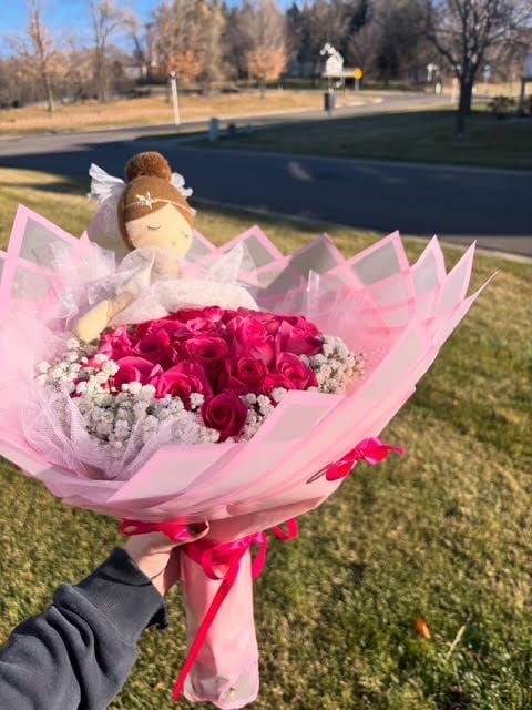 Hand holding a bouquet of red roses wrapped in pink tissue paper with an angel decoration on top, in a park setting
