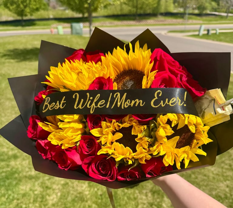 Hand holding bouquet of yellow sunflowers and red roses with Best Wife Mom Ever gold ribbon and black wrapper in park