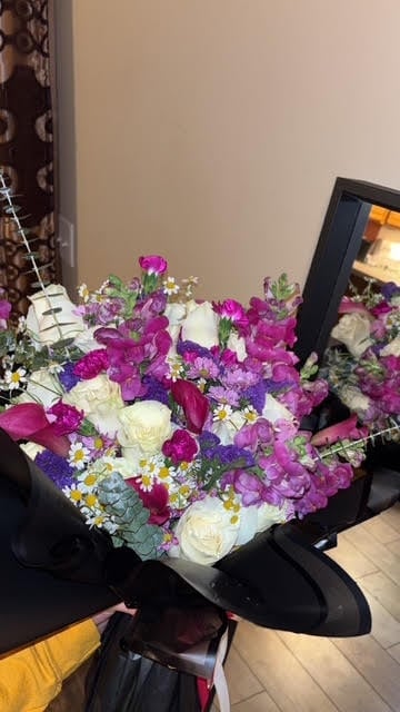 Large bouquet of mixed flowers including magenta roses, white roses, purple carnations, and yellow daisies with green foliage, wrapped in black paper